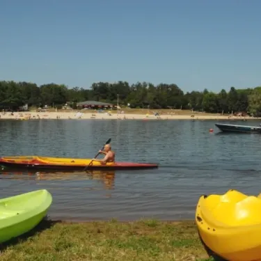 Un homme en kayak sur l'eau, avec des kayaks verts et jaunes à la rive. Camping Des Lacs.