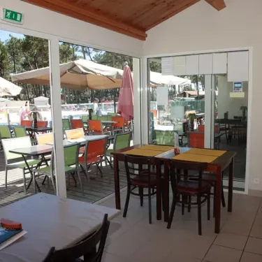 Intérieur du restaurant du Camping La Plage de Riez avec tables et chaises, vue sur la piscine extérieure.