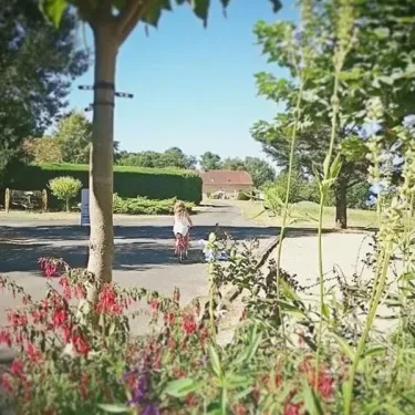Camping La Nouvelle Croze : enfant en vélo sur un chemin entouré de fleurs et d’arbres.