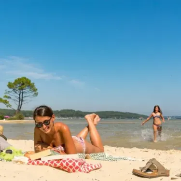 Femme lisant sur une plage avec enfants qui jouent dans l'eau au Camping Slow Village