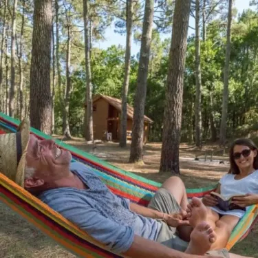 Couple dans une hamac dans la forêt, avec cabane en bois au loin à Camping Slow Village.