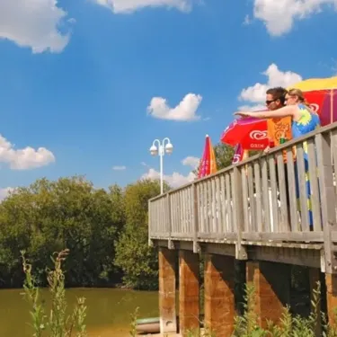 Deux personnes regardant le lac depuis un pont en bois au Camping L'Etang de Besse, sous un ciel bleu.