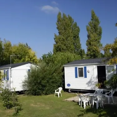 Camping Kerarno : deux mobil-homes blancs avec volets bleus entourés d'arbres et de chaises en plastique.