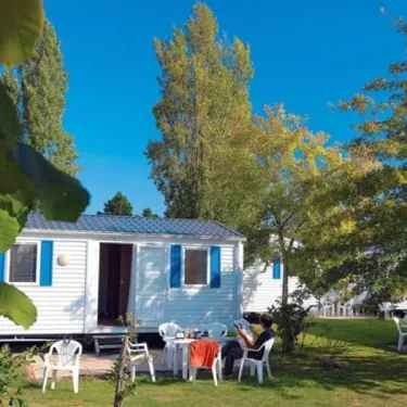 Camping Kerarno : mobil-home blanc avec volets bleus, table et chaises en plastique sur pelouse.