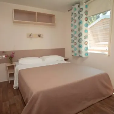 Chambre dans Camping Les Demoiselles : lit double, rideaux floraux et étagère murale