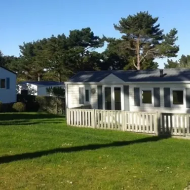 Camping Port de la Chaine : mobil-home blanc avec clôture en bois, pelouse verte et pins en arrière-plan.