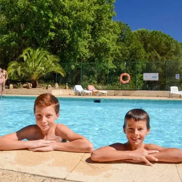 Deux enfants souriants à la bordure de la piscine dans un camping verdoyant