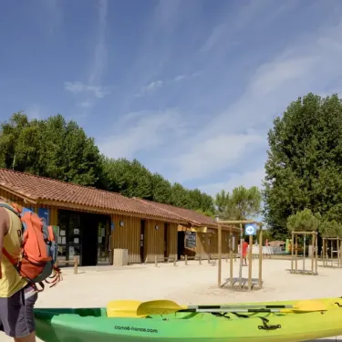 Personne portant un sac à dos près d'un kayak vert, devant une cabane en bois, Camping Les Ondines