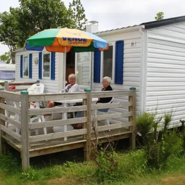 Camping La Plage : deux personnes assises sur une terrasse en bois devant une caravane blanche avec un parasol coloré.