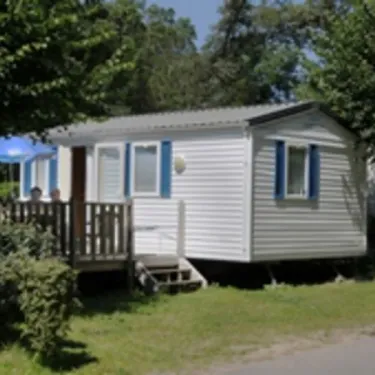 Camping Du Kérou : caravane blanche avec volets bleus, devant un jardin et une terrasse ombragée.