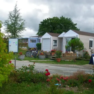 Caravane beige avec vélos garés devant, jardin fleuri et panneau d'information au Camping Le Pontis.