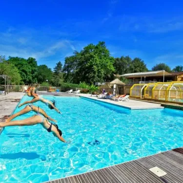 Trois femmes sautent dans la piscine du Camping Des Chênes Verts sous un ciel bleu.