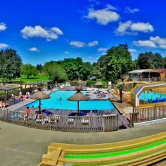 Vue panoramique du camping avec piscine, parasols et personnes détendues sous un ciel bleu.