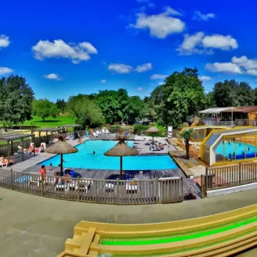 Vue panoramique du camping avec piscine, parasols et personnes détendues sous un ciel bleu.