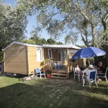 Camping Le Vorlen : mobil-home en bois avec table et chaises sous un parasol bleu.
