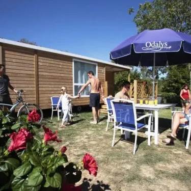 Camping Le Vorlen : famille autour d'une table sous un parasol Odalys, près d'un mobil-home en bois.