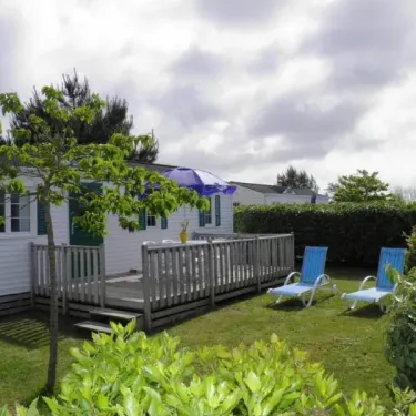Camping Saint Martin : mobil-home blanc avec volets verts, terrasse, chaises longues et parasol bleu.