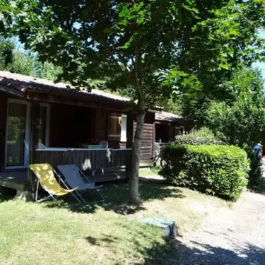 Camping Lé Moulis : cabane en bois avec terrasse et chaise longue jaune sous un arbre ombragé.