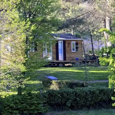 Camping Le Bosquet 12 : cabane en bois avec toit en tuiles, entourée d'arbres et de haies vertes.