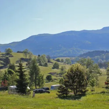 Village de camping avec caravanes et voitures sur une colline verdoyante, montagnes en fond.