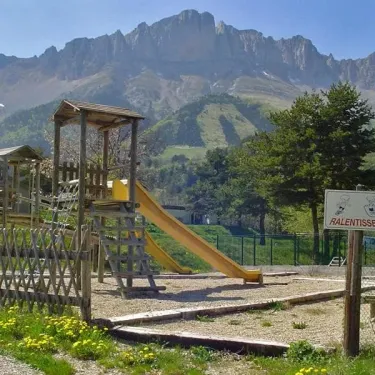 Piste de jeux en bois avec toboggan jaune au Camping Les 4 Saisons, montagnes en arrière-plan.