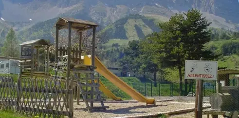 Piste de jeux en bois avec toboggan jaune au Camping Les 4 Saisons, montagnes en arrière-plan.