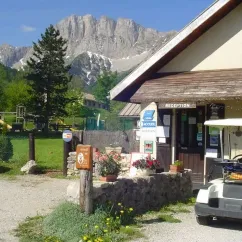 Entrée du camping avec reception et voiturette blanche devant un bâtiment en pierre, montagnes à l'horizon.
