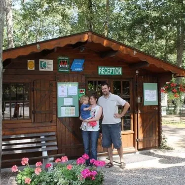 Une famille devant la réception du camping Les Acacias - 37, avec des fleurs et un petit banc.
