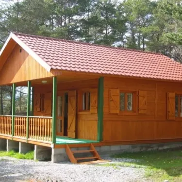 Cabane en bois avec toit rouge et balcon vert au Camping Le Champ Long, entourée de pins.
