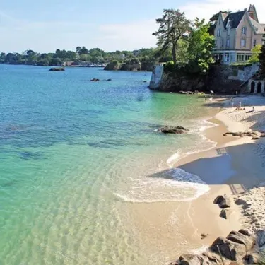 Camping Kerscolper : plage de sable fin bordée d'océan turquoise et de rochers, avec un bâtiment au loin.
