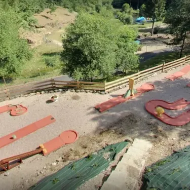 Piste de mini-golf en terre rouge entourée de verdure, Camping Les Trois Sources.