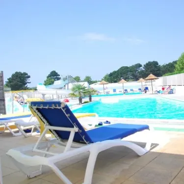 Lits longues bleus sur une terrasse ensoleillée près d'une piscine avec parasols et arbres, au Camping Du Domaine de L'Auzance.